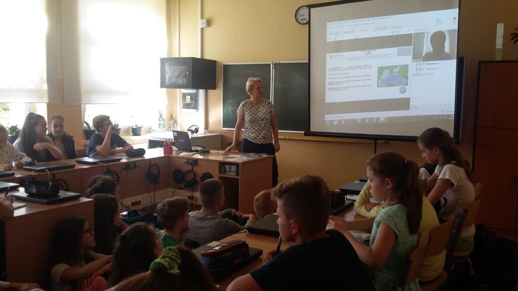 Klasa szkolna, uczniowie siedzą w ławkach i na podłodze a nauczycielka stoi przy biurku zwrócona do tablicy (ekran rzutnika) i omawia temat związany z zajęciami